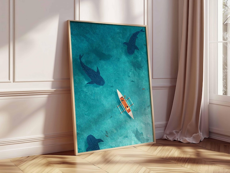 May include: A framed art print featuring an aerial view of a turquoise ocean with dark blue whale shark silhouettes. A small white boat with orange figures is in the center. The artwork is displayed on a wooden floor next to a white wall and a beige curtain.