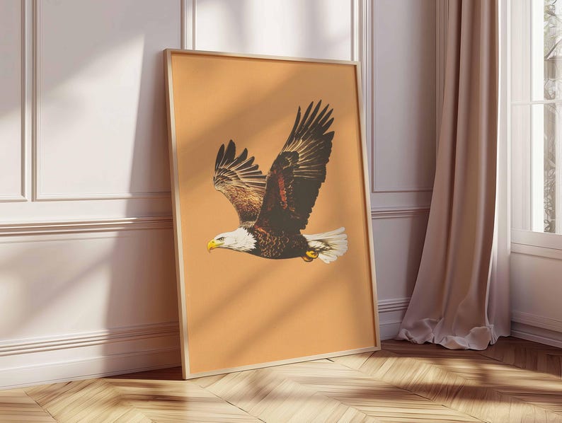May include: A framed print featuring a soaring bald eagle with outstretched wings. The eagle has brown and white feathers, a yellow beak, and is set against a peach-colored background. The frame is a light wood color, and the print is leaning against a white wall.