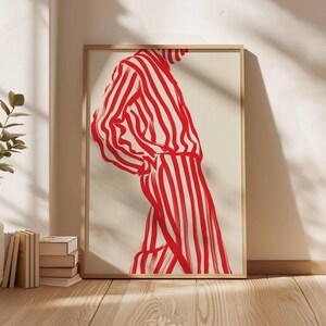 Puede incluir: Impresi&oacute;n art&iacute;stica enmarcada que presenta una figura con un atuendo a rayas rojas y blancas. La obra de arte es una ilustraci&oacute;n estilizada con trazos audaces, sobre un fondo neutro. La impresi&oacute;n se muestra en un marco de madera clara.