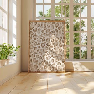 May include: A framed artwork featuring a leopard print design in shades of brown and beige. The artwork is displayed in a room with natural light from a large window. The room has light-colored wooden floors and a neutral wall color.