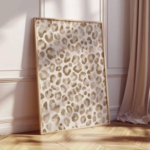 May include: A framed art print featuring a leopard print pattern in shades of beige and brown. The artwork is displayed in a light-colored wooden frame and leans against a white wall and wooden floor, with a curtain and window in the background.