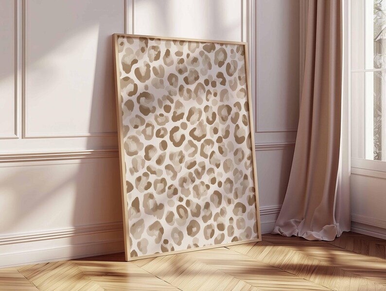 May include: A framed art print featuring a leopard print pattern in shades of beige and brown. The artwork is displayed in a light-colored wooden frame and leans against a white wall and wooden floor, with a curtain and window in the background.