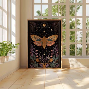 May include: A framed art print featuring a detailed illustration of a moth with intricate wing patterns in shades of orange, brown, and gold. The artwork is set against a black background with celestial elements and floral accents. The print is displayed in a room with natural light.