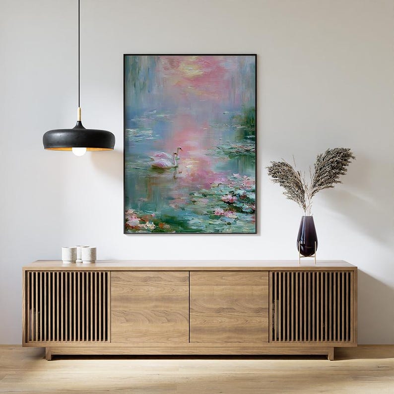 May include: An oil painting in a black frame depicts a serene water scene with two white swans. The artwork features a palette of pink, blue, and green, with lily pads and a soft, impressionistic style. The painting hangs above a wooden cabinet.