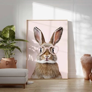 K&ouml;nnte beinhalten: Gerahmter Druck mit einem Hasen, der eine rosa herzf&ouml;rmige Brille vor einem hellrosa Hintergrund tr&auml;gt. Der Hase hat braunes und wei&szlig;es Fell und lange Ohren. Das Kunstwerk wird in einem Raum mit einer Pflanze, einem Hocker und dekorativen Vasen ausgestellt.
