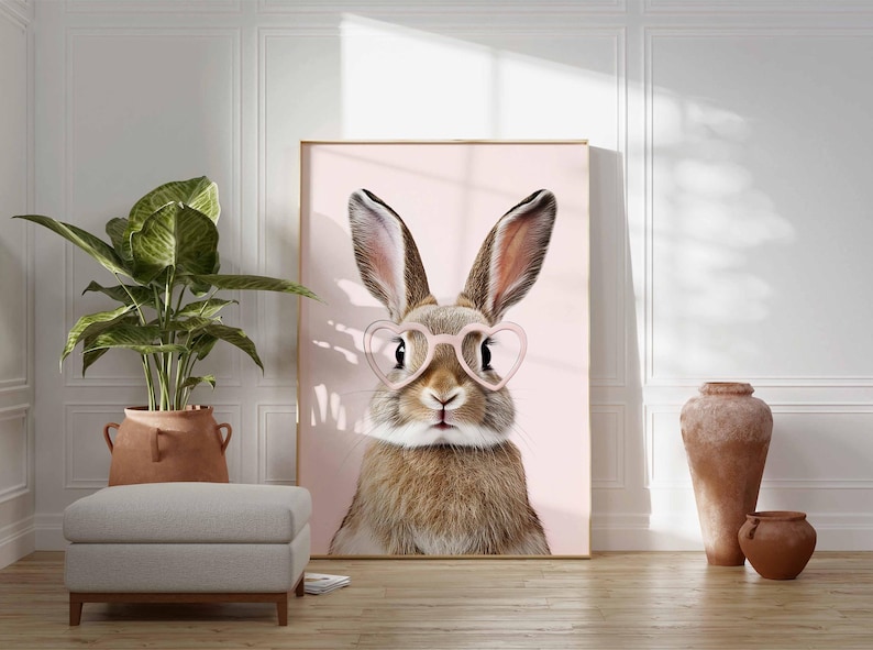K&ouml;nnte beinhalten: Gerahmter Druck mit einem Hasen, der eine rosa herzf&ouml;rmige Brille vor einem hellrosa Hintergrund tr&auml;gt. Der Hase hat braunes und wei&szlig;es Fell und lange Ohren. Das Kunstwerk wird in einem Raum mit einer Pflanze, einem Hocker und dekorativen Vasen ausgestellt.
