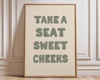 May include: A framed print with the text "TAKE A SEAT SWEET CHEEKS" in green, stylized lettering. The print has a cream-colored background and a wooden frame. The artwork is leaning against a white wall and a wooden floor.