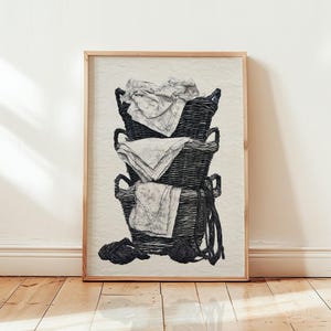 May include: A framed black and white line art print depicting a vertical stack of three woven wicker baskets. Each basket contains a folded piece of fabric, and a length of rope hangs down from the side of the bottom basket onto the base.