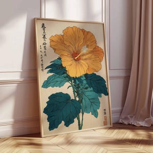 May include: A framed botanical print of a large yellow hibiscus flower with a red centre and green leaves. The artwork includes Japanese text and is set against a beige background. The frame is light wood, and the print leans against a white wall.