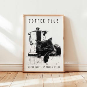 May include: A framed monochrome print showing a black cat lounging near a coffee maker. The text "COFFEE CLUB" is above the image, with "WHERE EVERY CUP TELLS A STORY" below. The frame is made of light-colored wood.