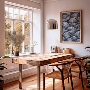 May include: A light-filled room with a wooden desk and two chairs. A framed artwork with a blue and white wave pattern hangs on the wall. A large window offers a view of trees. A silver pendant light hangs above the desk.
