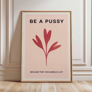 Op de afbeelding: Ingelijste kunstdruk met de tekst "BE A PUSSY" boven een rood bloemmotief. Onder het ontwerp staat de tekst "BECAUSE THEY CAN HANDLE A LOT". Het kunstwerk staat tegen een lichtroze achtergrond.