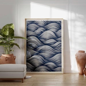 May include: A framed art print featuring a navy blue and white wave pattern. The artwork is displayed in a light-colored wooden frame, leaning against a white wall. The room also includes a potted plant, a gray ottoman, and terracotta vases.
