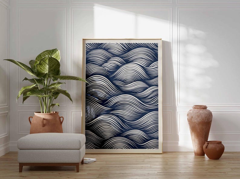 May include: A framed art print featuring a navy blue and white wave pattern. The artwork is displayed in a light-colored wooden frame, leaning against a white wall. The room also includes a potted plant, a gray ottoman, and terracotta vases.