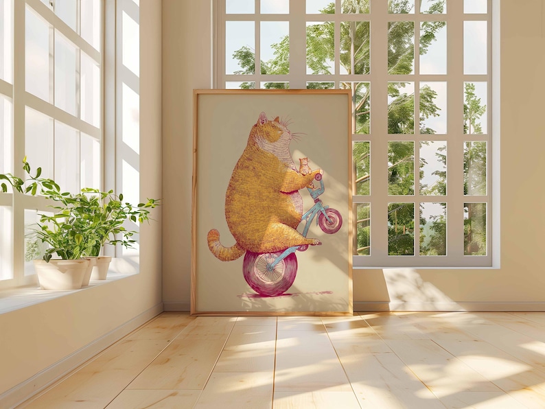K&ouml;nnte beinhalten: Ein gerahmter Kunstdruck zeigt eine gro&szlig;e orange Katze auf einem kleinen blauen Fahrrad mit einem rosa Rad. Die Katze l&auml;chelt. Das Kunstwerk wird in einem Raum mit Fenster und Pflanzen ausgestellt.