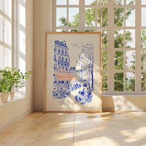 May include: A framed art print depicting a Parisian cafe scene in blue and orange hues. The artwork features buildings, trees, and people seated at tables. The print is displayed in a room with a large window and potted plants.