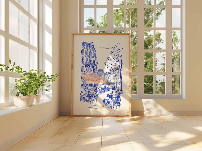 May include: A framed art print depicting a Parisian cafe scene in blue and orange hues. The artwork features buildings, trees, and people seated at tables. The print is displayed in a room with a large window and potted plants.