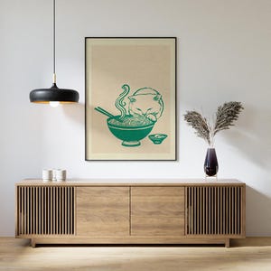 May include: A framed wall art print featuring a green line drawing of a cat eating noodles from a bowl with chopsticks, set against a light beige background. The artwork is displayed in a thin black frame hanging on a plain white wall.