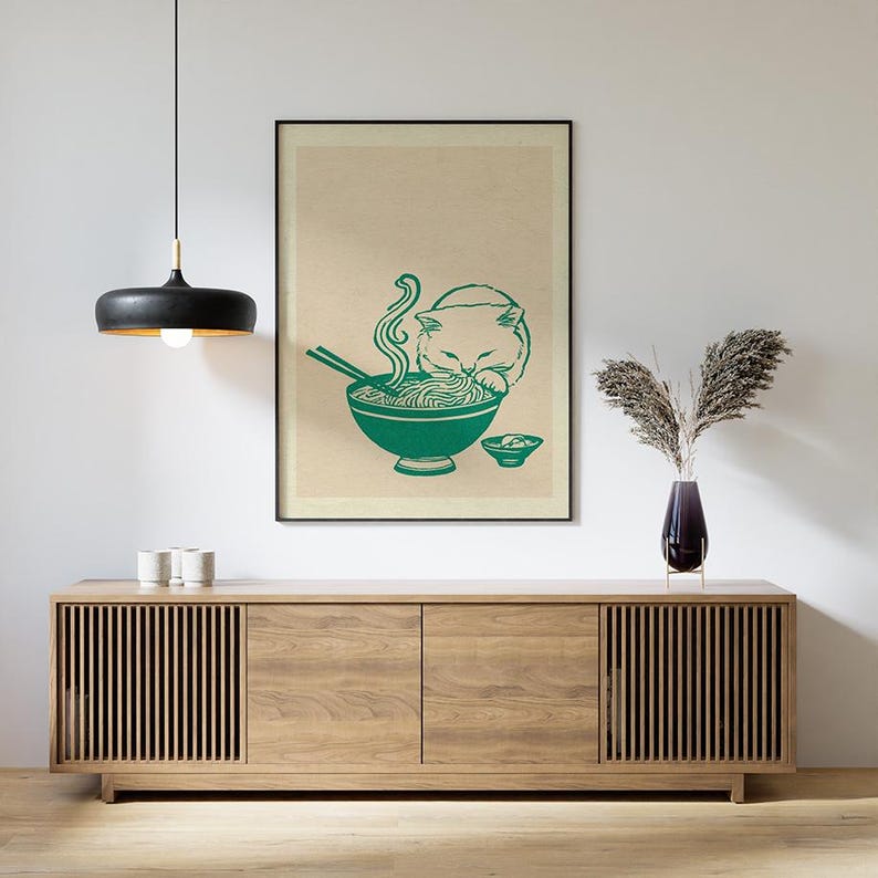 May include: A framed wall art print featuring a green line drawing of a cat eating noodles from a bowl with chopsticks, set against a light beige background. The artwork is displayed in a thin black frame hanging on a plain white wall.
