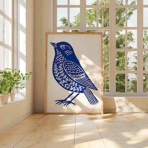 May include: A framed art print featuring a blue bird illustration with white leaf-like patterns. The artwork is displayed in a bright room with a window and potted plants. The bird is standing, facing left.