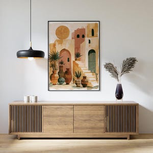 Peut inclure: Une impression encadr&eacute;e &agrave; l'aquarelle repr&eacute;sente un village m&eacute;diterran&eacute;en avec des b&acirc;timents en terre cuite, des arches et des escaliers. L'&oelig;uvre est au-dessus d'un meuble en bois avec une suspension noire et un vase de plantes s&eacute;ch&eacute;es.