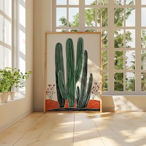 May include: A framed art print featuring a stylized illustration of green cacti against a cream background. The artwork includes a red and black base with small red floral accents. The print is displayed in a bright room with a window.