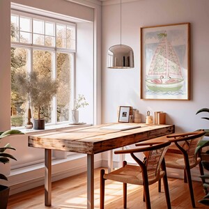 May include: A light-filled room with a wooden desk and two chairs. A framed artwork of a sailboat with pink flamingos hangs on the wall. A large window offers a view of trees. A chrome pendant light hangs above the desk.