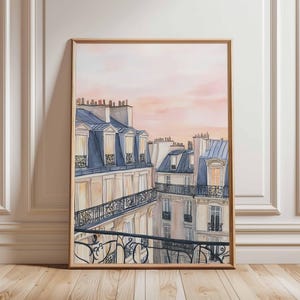 May include: Watercolor painting of Parisian rooftops with blue-gray mansard roofs, ornate balconies, and cream-colored walls. The artwork is framed in a light brown wooden frame, set against a soft pink and peach sky.