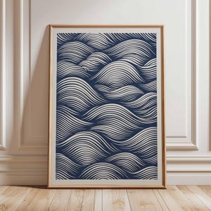 May include: A framed art print featuring a navy blue and white wave pattern. The artwork is displayed in a light brown wooden frame, leaning against a white wall and wooden floor.