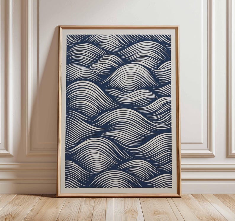 May include: A framed art print featuring a navy blue and white wave pattern. The artwork is displayed in a light brown wooden frame, leaning against a white wall and wooden floor.