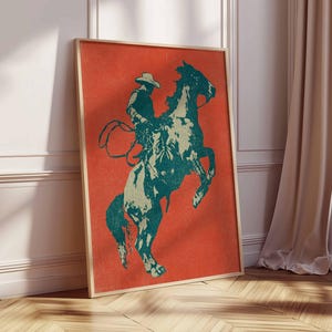 May include: A framed art print featuring a cowboy on a rearing horse. The artwork uses a teal and off-white color palette against a vibrant orange background. The print is displayed in a room with a wooden floor and a beige curtain.