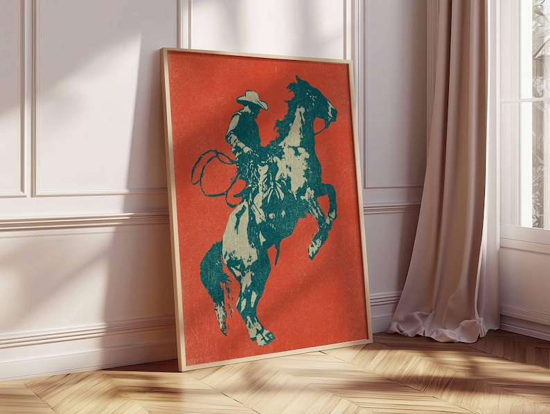 May include: A framed art print featuring a cowboy on a rearing horse. The artwork uses a teal and off-white color palette against a vibrant orange background. The print is displayed in a room with a wooden floor and a beige curtain.