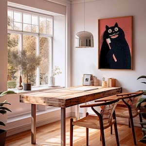 Op de afbeelding: Een rustiek houten bureau met metalen poten en twee stoelen met vlechtwerk. Een ingelijst kunstwerk van een zwarte kat die zijn tanden poetst, hangt aan de muur. De kamer heeft een groot raam en planten.