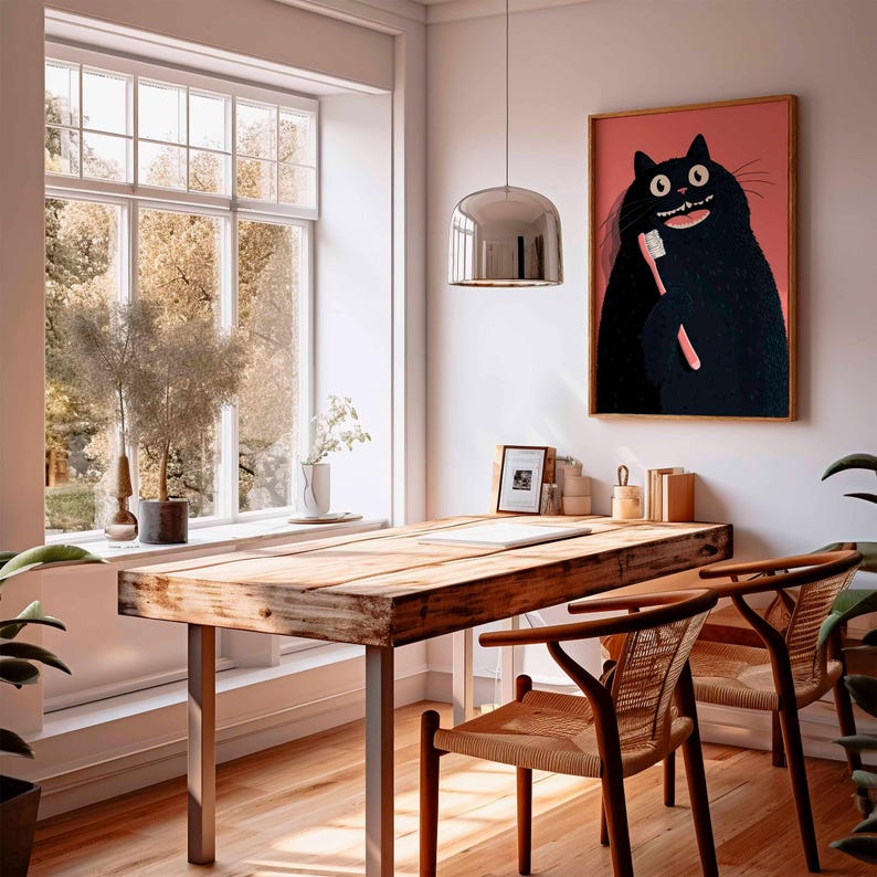 Op de afbeelding: Een rustiek houten bureau met metalen poten en twee stoelen met vlechtwerk. Een ingelijst kunstwerk van een zwarte kat die zijn tanden poetst, hangt aan de muur. De kamer heeft een groot raam en planten.