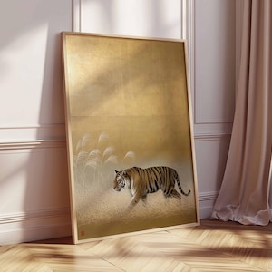 May include: Framed art print featuring a tiger walking through tall grass. The artwork has a gold background with a sepia-toned tiger and grass. The frame is a light wood color. The artwork is leaning against a white wall and a beige curtain.