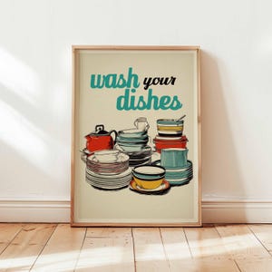 May include: A framed print featuring a retro illustration of stacked dishes, cups, and a teapot in red, teal, yellow, and white. The words "wash your dishes" are written in teal and black above the dishes.