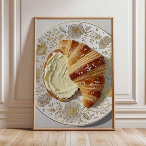 Könnte beinhalten: Gerahmter Kunstdruck mit einem Croissant und einer Scheibe Brot mit Butter auf einem dekorativen Teller. Der Teller hat einen weißen Hintergrund mit goldenen Blumenmustern. Das Croissant ist goldbraun mit Sesamkörnern.