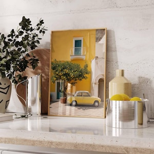 May include: A framed print depicts a yellow building with a small balcony and a yellow car parked in front. A lemon tree is in the center. The print is displayed on a countertop with decorative items, including a vase and a metal bowl.