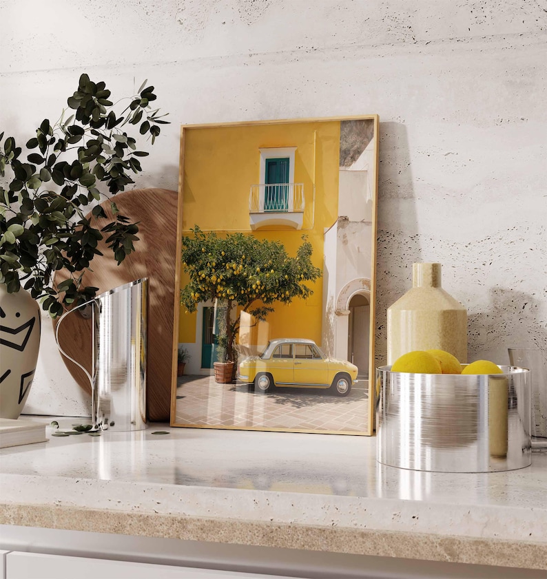 May include: A framed print depicts a yellow building with a small balcony and a yellow car parked in front. A lemon tree is in the center. The print is displayed on a countertop with decorative items, including a vase and a metal bowl.
