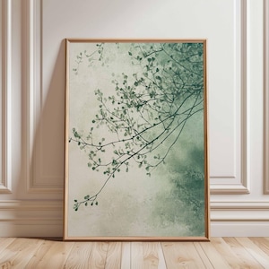 Op de afbeelding: Een ingelijste kunstdruk met een aquarelachtige weergave van boomtakken met kleine groene bladeren. Het kunstwerk heeft een zacht, gedempt kleurenpalet van groen en beige, en is ingelijst in een lichtbruine houten lijst.