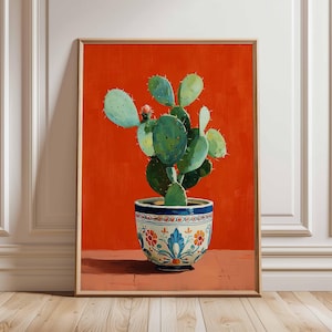 Op de afbeelding: Een ingelijst schilderij van een groene cactus in een decoratieve pot. De pot is wit met blauwe en oranje bloemmotieven. De achtergrond is effen oranje. Het kunstwerk wordt tentoongesteld in een kamer met witte lambrisering.