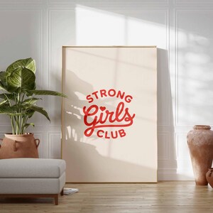 Pu&ograve; includere: Una grande stampa incorniciata con le parole "STRONG Girls CLUB" in corsivo rosso. La stampa &egrave; appoggiata a una parete bianca. Una pianta in vaso, un pouf grigio e due vasi in terracotta sono in primo piano.