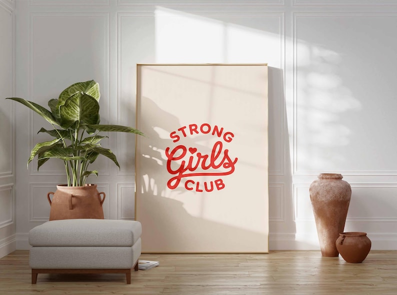 Pu&ograve; includere: Una grande stampa incorniciata con le parole "STRONG Girls CLUB" in corsivo rosso. La stampa &egrave; appoggiata a una parete bianca. Una pianta in vaso, un pouf grigio e due vasi in terracotta sono in primo piano.