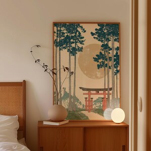 May include: A framed art print depicting a Japanese landscape with tall trees, a red torii gate, and a large, pale sun. The artwork is in a wooden frame and hangs above a wooden cabinet. A round vase and a small lamp are on the cabinet.
