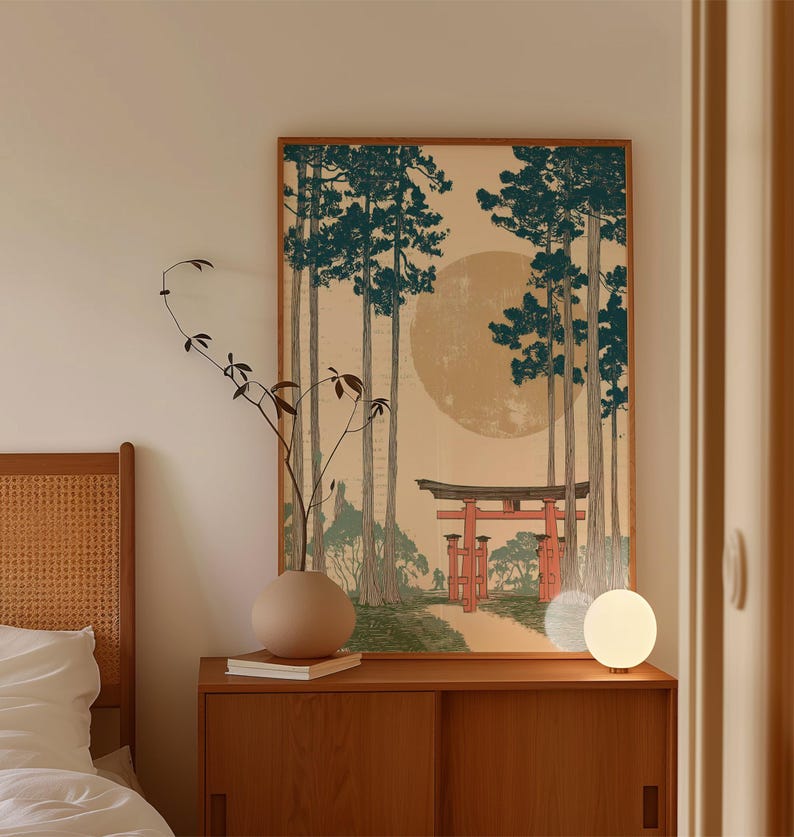 May include: A framed art print depicting a Japanese landscape with tall trees, a red torii gate, and a large, pale sun. The artwork is in a wooden frame and hangs above a wooden cabinet. A round vase and a small lamp are on the cabinet.