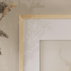 May include: A framed art print with a deer silhouette in a soft, neutral colour scheme. The artwork is set within a light wood frame, creating a minimalist and natural aesthetic. The background has a subtle tree pattern.