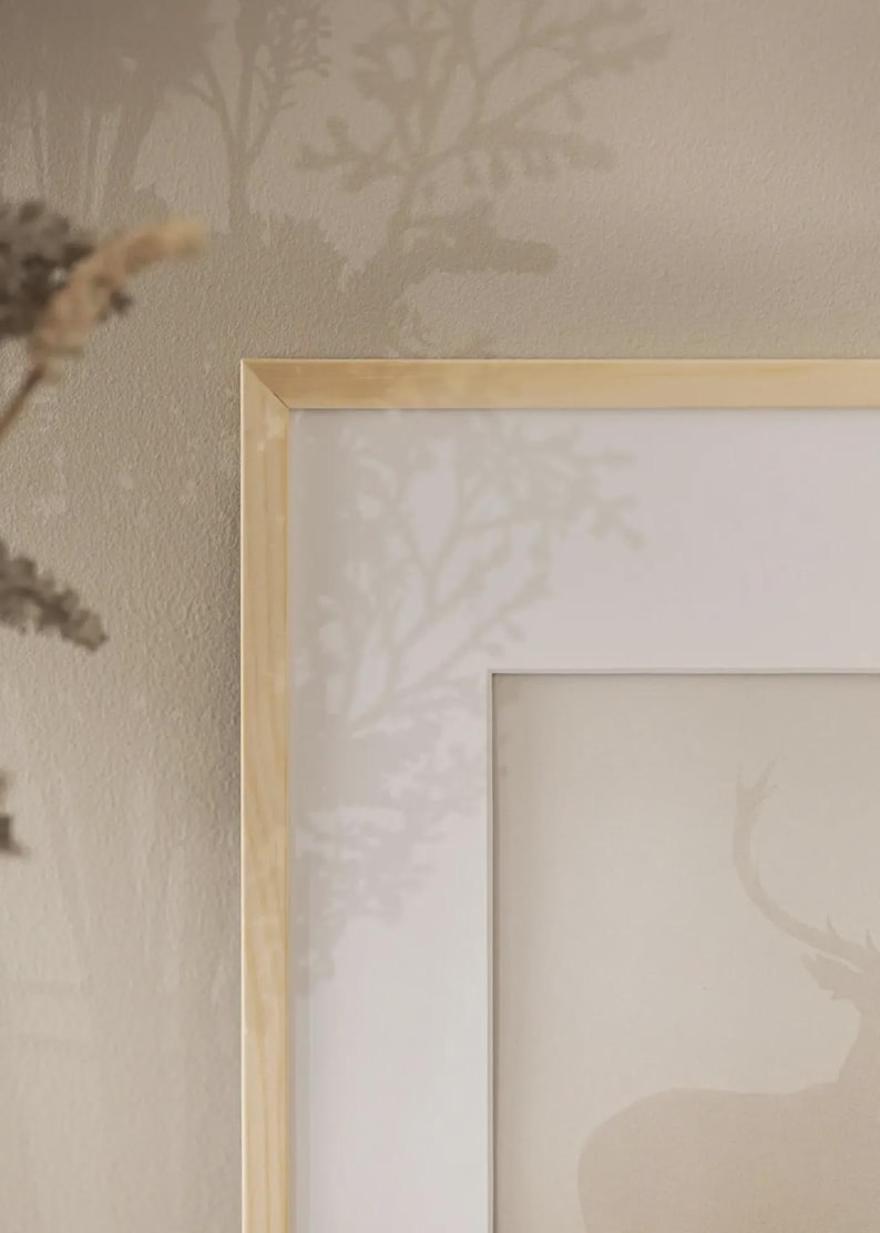 May include: A framed art print with a deer silhouette in a soft, neutral colour scheme. The artwork is set within a light wood frame, creating a minimalist and natural aesthetic. The background has a subtle tree pattern.