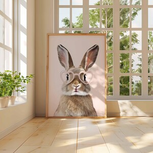 K&ouml;nnte beinhalten: Ein gerahmter Druck mit einem Hasen, der eine herzf&ouml;rmige Brille tr&auml;gt. Der Hase hat braunes und hellbraunes Fell, vor einem hellrosa Hintergrund. Der Druck wird in einem Raum mit nat&uuml;rlichem Licht und Holzboden ausgestellt.