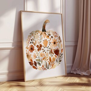 May include: A framed watercolor print featuring a pumpkin shape composed of autumn-colored flowers and leaves. The artwork is in a wooden frame and leans against a white wall and a wooden floor.