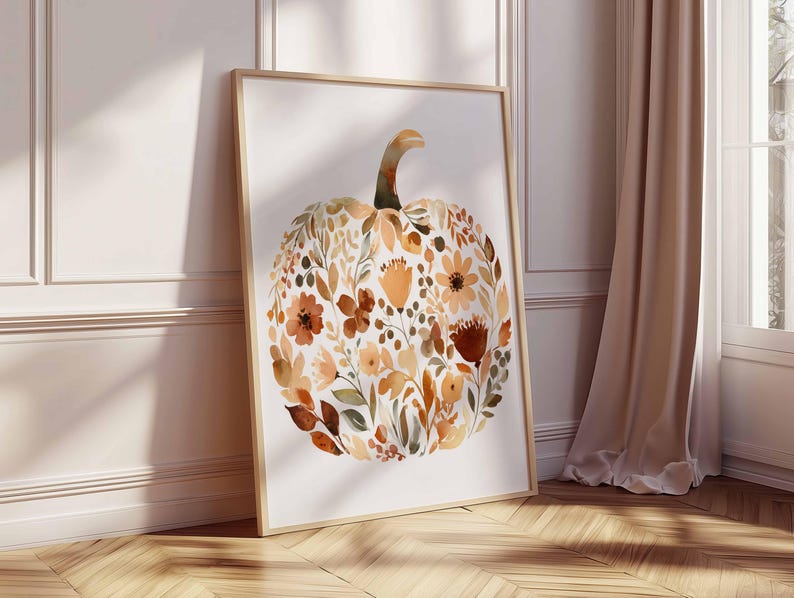 May include: A framed watercolor print featuring a pumpkin shape composed of autumn-colored flowers and leaves. The artwork is in a wooden frame and leans against a white wall and a wooden floor.